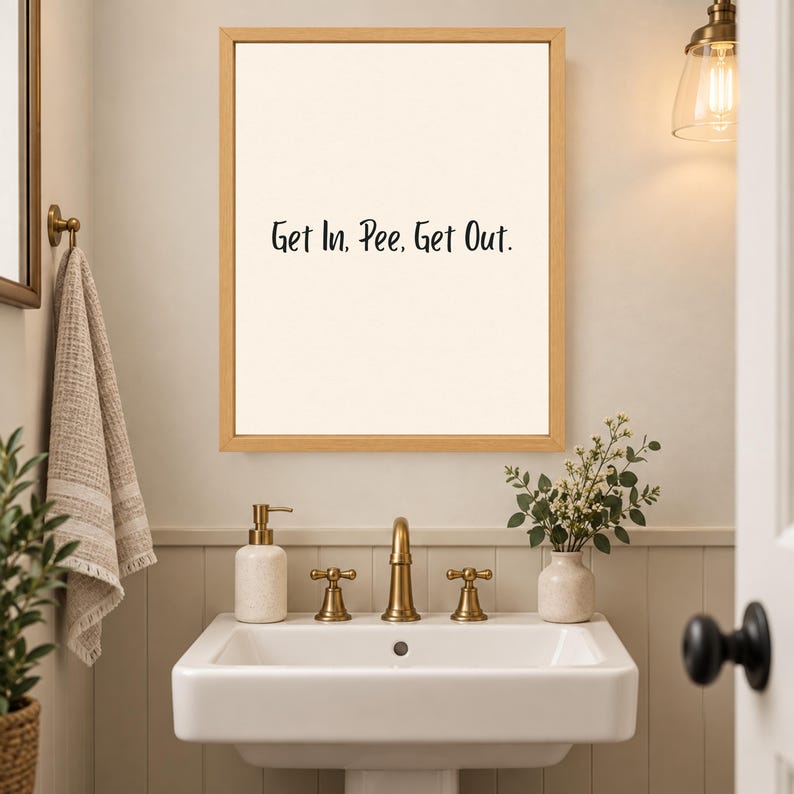 May include: A rectangular wall art piece in a light wood frame featuring a cream-colored background with the black text Get In, Pee, Get Out. The artwork is displayed above a white bathroom sink with brass fixtures and a small potted plant.