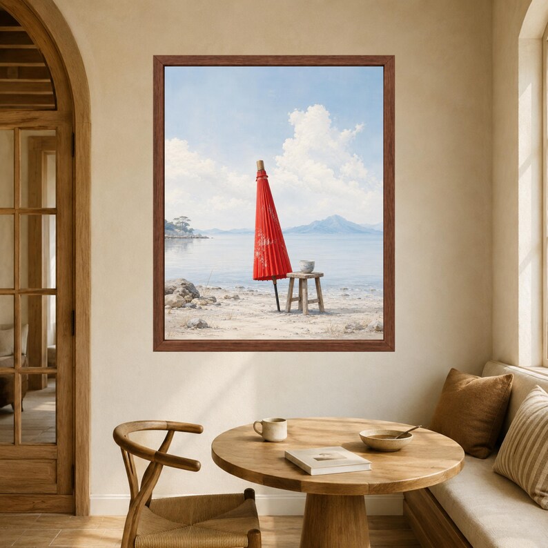 May include: A framed wall art print depicting a bright red paper parasol standing upright on a sandy beach next to a small wooden stool. The scene features a calm body of water, distant mountains, and a light blue sky with white clouds.