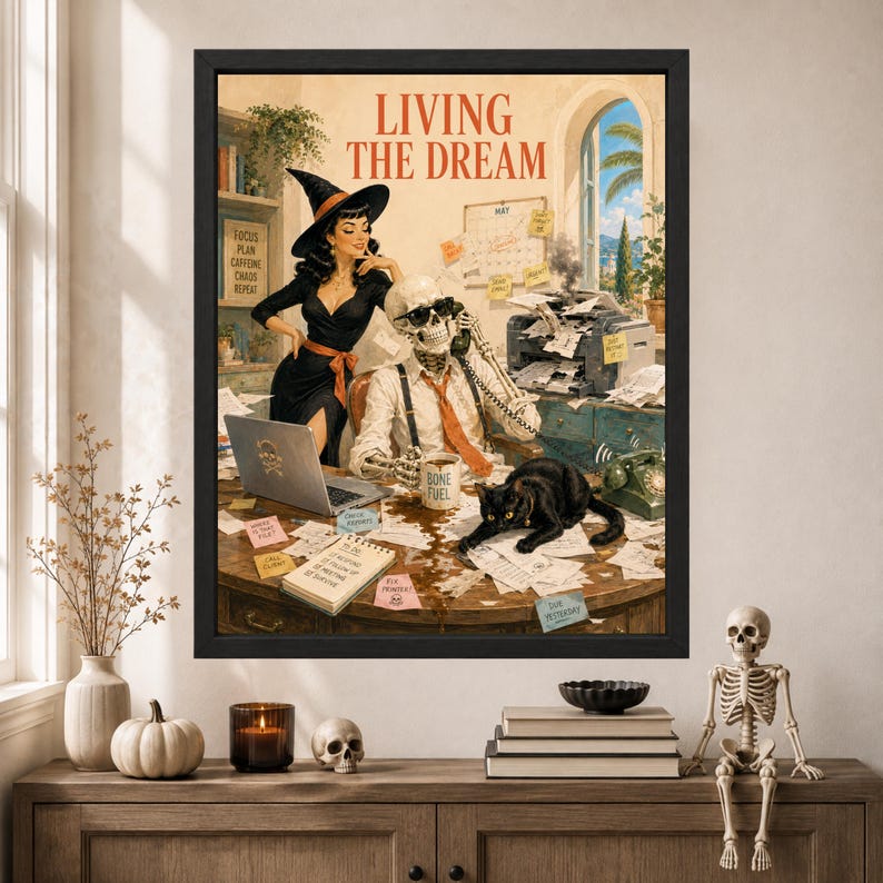 May include: A framed wall art print titled LIVING THE DREAM featuring a witch in a black dress and a skeleton in a white shirt and orange tie at a desk. A black cat sits on a cluttered desk with a laptop, printer, and sticky notes.