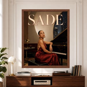 Framed Sade wall art featuring a woman in a deep red satin dress seated beside a black grand piano in a warm late-night lounge with large cream SADE lettering above.