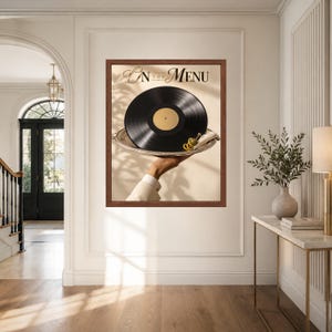 May include: A framed wall art print titled On The Menu featuring a black vinyl record served on a silver platter. A hand in a white sleeve holds the tray, which includes a small green olive garnish. The background is a light beige wall.