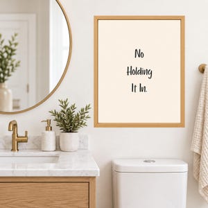 May include: A rectangular wall art print in a light wood frame featuring the black text No Holding It In centered on a cream background. The artwork is displayed on a wall above a white toilet, positioned near a bathroom vanity and mirror.