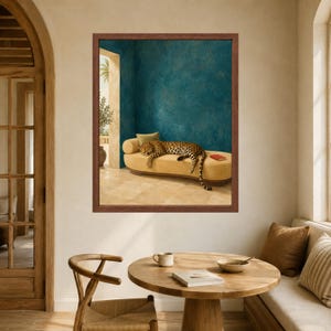 May include: A framed wall art print depicting a leopard resting on a long, curved, mustard-yellow upholstered bench. The scene is set against a textured teal wall and a light-colored stone floor, with a small red book resting on the bench.