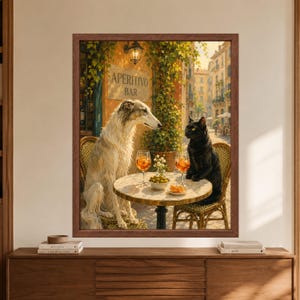 Framed whimsical animal wall art showing a long-haired greyhound and black cat sharing orange aperitivo drinks and olives at a small Italian cafe table on a sunlit European street.