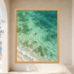 May include: A framed wall art print featuring an aerial view of clear turquoise ocean water with a sandy shoreline and two small figures swimming in the center. The water displays light-refracting patterns and visible rocky seabed textures.