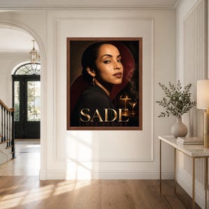 K&ouml;nnte beinhalten: Ein gerahmter Kunstdruck mit einem Portr&auml;t von Sade und dem Text SADE LOVE DELUXE. Das Bild zeigt eine Person in einem dunklen Blazer vor einem dunklen Hintergrund mit einer leuchtenden Lampe und einem Cocktailglas. Der Rahmen ist aus mittelbraunem Holz.
