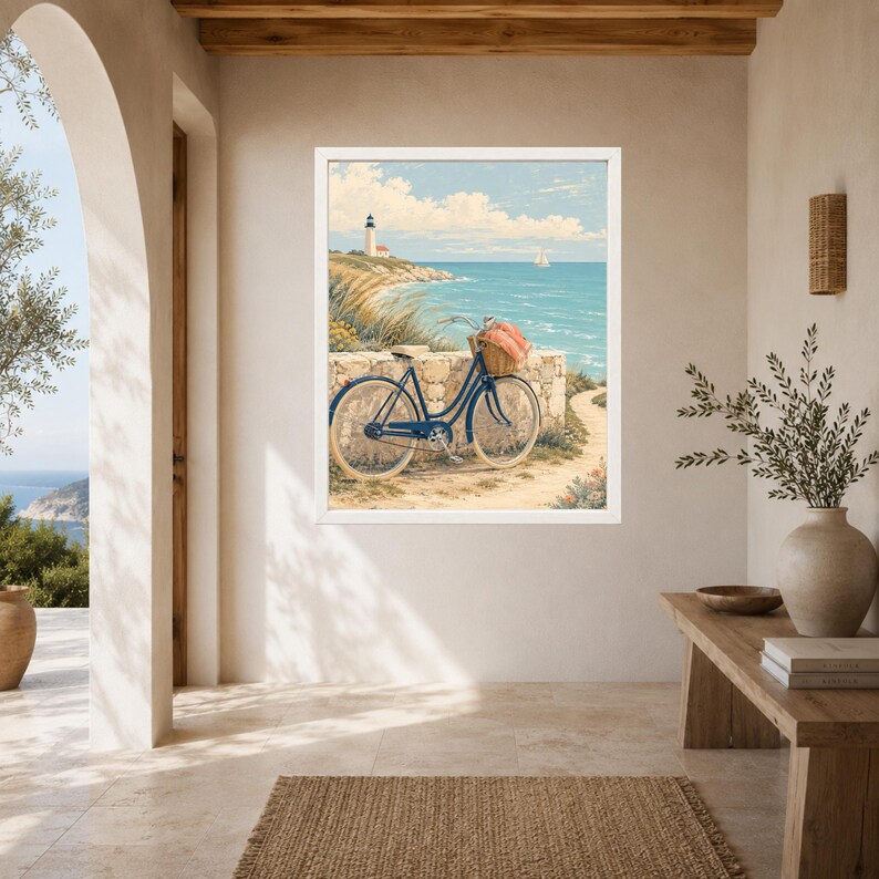 Op de afbeelding: Een ingelijste kunstdruk van een kustlandschap met een blauwe fiets geparkeerd bij een stenen muur. De fiets heeft een rieten mand aan het stuur met een roze doek erin. Op de achtergrond staat een vuurtoren op een klif aan de zee.