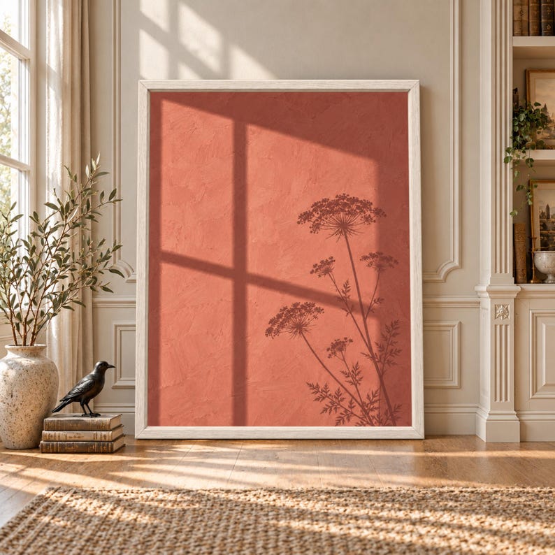 Framed terracotta botanical wall art showing delicate flower silhouettes and a large window shadow across a textured warm coral wall in soft afternoon sunlight.