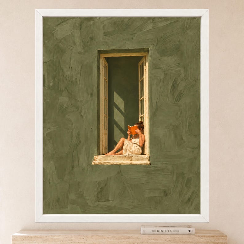 May include: A framed wall art print featuring a textured olive green wall with a central open window. A figure sits on the window ledge reading an orange book while wearing a light-colored dress. The artwork is enclosed in a simple white frame.