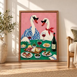 Framed whimsical swan wall art showing two dressed swans sharing red wine, cheese, olives and appetizers at a green dinner table against a pink background.