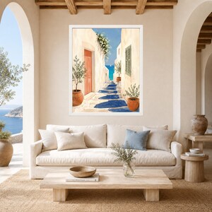 May include: A framed wall art print depicting a narrow Mediterranean alleyway with cream-colored buildings, a bright orange door, potted green plants, and a path leading toward a vibrant blue sea under a clear sky with deep blue shadows.