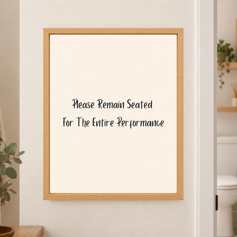 Framed minimalist bathroom wall art with the handwritten black phrase “Please Remain Seated for the Entire Performance” centered on a warm off-white background in a light wood frame beside a bathroom doorway.