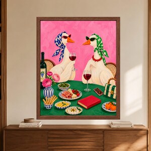 May include: A framed wall art print featuring two white geese wearing headscarves and sunglasses, sitting at a green table with wine, breadsticks, fruit, and a red book. The background is a solid bright pink, and the geese are positioned in chairs.