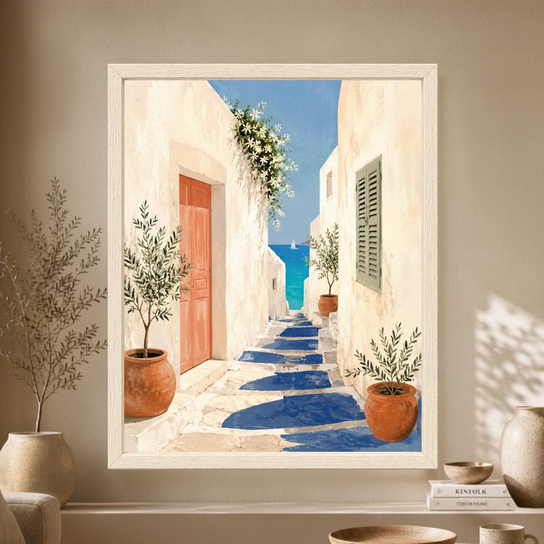 Framed Mediterranean coastal wall art showing a whitewashed stone alley with a coral door, green shutters, potted olive trees and steps leading toward a bright blue sea with a small sailboat.