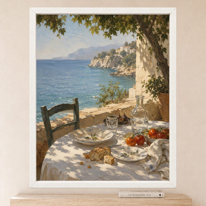 May include: A framed wall art print depicting a coastal scene with a table set for a meal. The table features a white tablecloth, a glass carafe, a plate with bread, a bowl of tomatoes, and a glass. A wooden chair sits by the sea under a tree.