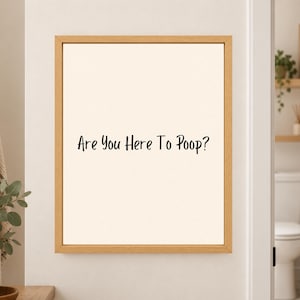 Framed minimalist bathroom wall art with the black handwritten phrase “Are You Here To Poop?” centered on a warm off-white background in a light wood frame beside a bathroom doorway.