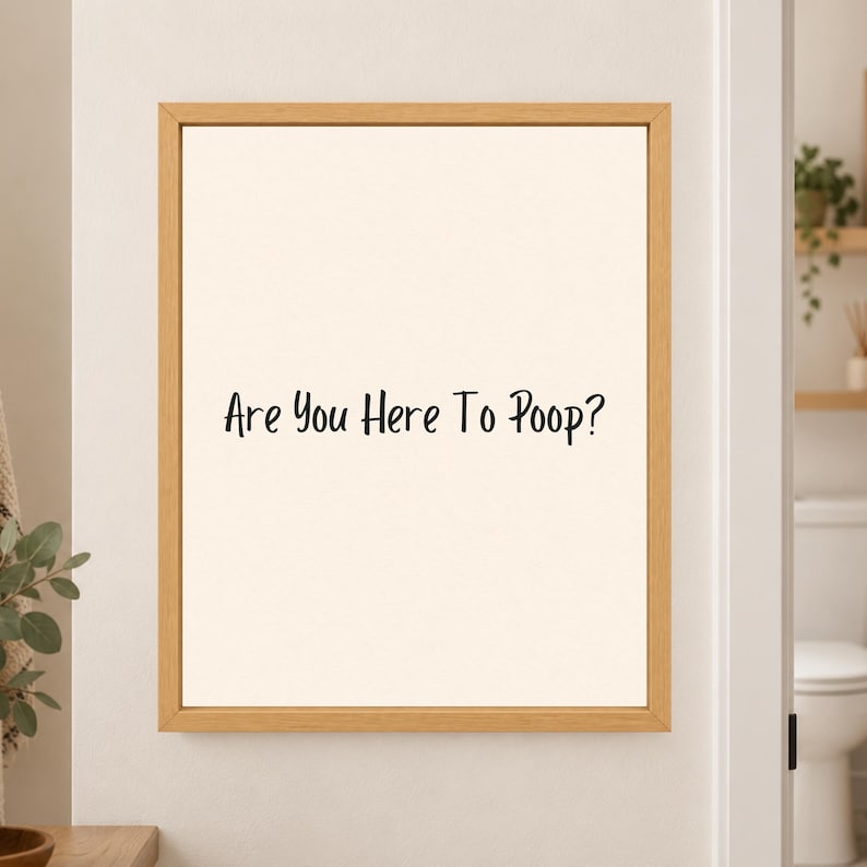 Framed minimalist bathroom wall art with the black handwritten phrase “Are You Here To Poop?” centered on a warm off-white background in a light wood frame beside a bathroom doorway.