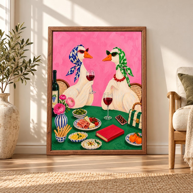 Framed pink duck wine party wall art showing two stylish white ducks in sunglasses and patterned headscarves sharing red wine, cheese, olives and snacks at a green table.