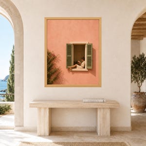 May include: A rectangular wall art print in a light wood frame featuring a peach-colored stucco wall with an open window. Green shutters flank the window, where a person sits reading a book. A climbing vine grows along the left side of the wall.