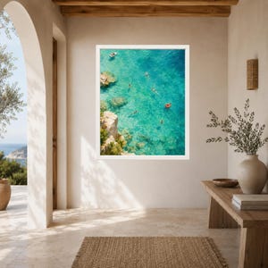 Puede incluir: Una impresi&oacute;n art&iacute;stica enmarcada que muestra una vista a&eacute;rea de agua de mar turquesa con acantilados rocosos, varios nadadores y un peque&ntilde;o barco blanco. Un flotador circular rojo brillante es visible en el agua, contrastando con el mar.