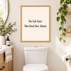 May include: A rectangular wall art print in a light wood frame featuring a cream-colored background with the black text "You Look Good Now Wash Your Hands" centered in the middle of the frame, displayed above a white toilet in a bathroom.