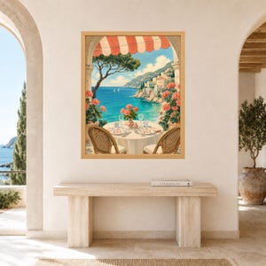 May include: A framed wall art print depicting a coastal Mediterranean scene viewed from a terrace. The image features a round table set for two with wicker chairs, pink geraniums, a red and white striped awning, and a blue sea with cliffs.