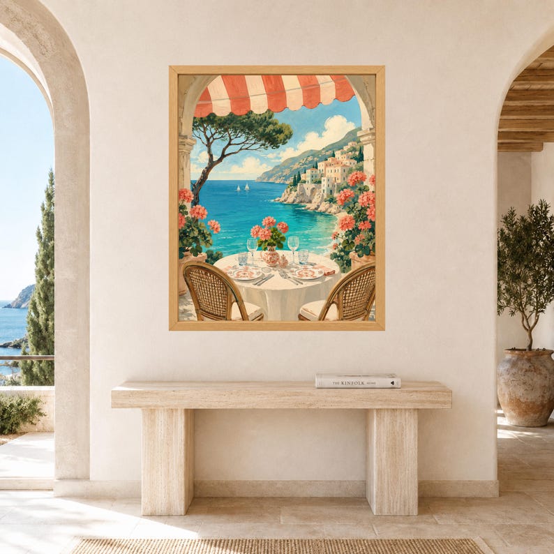 May include: A framed wall art print depicting a coastal Mediterranean scene viewed from a terrace. The image features a round table set for two with wicker chairs, pink geraniums, a red and white striped awning, and a blue sea with cliffs.