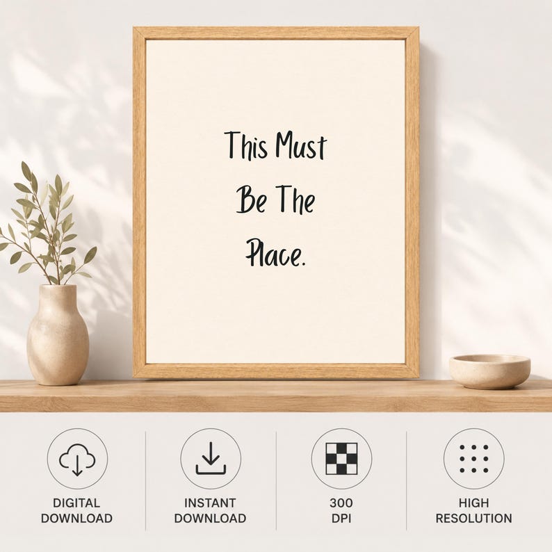 May include: A framed wall art print featuring the black text This Must Be The Place. centered on a cream-colored background. The print is housed in a light-toned wooden frame. Below the frame, four circular icons indicate digital download features.