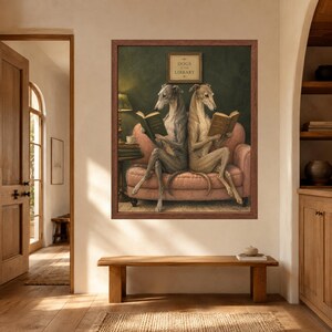 May include: A framed wall art print featuring two greyhounds sitting back-to-back on a dusty pink sofa, each reading a book. Above them, a small sign reads DOGS AT THE LIBRARY. The background is a dark green wall with a lamp on a side table.