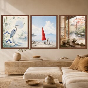 May include: A set of three framed wall art prints. The left print shows a blue heron near bamboo and irises. The center print depicts a red parasol on a beach. The right print shows a wooden table with a tea set and an open book by a window.