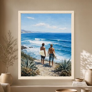 Framed surfer wall art showing two surfers holding white surfboards on a rocky coastal path while looking over blue ocean waves, distant cliffs and agave plants.