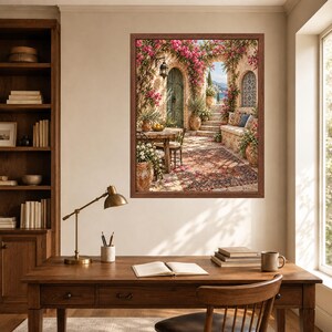 May include: A framed wall art print depicting a Mediterranean patio scene with a stone archway, vibrant pink bougainvillea flowers, a wooden table, chairs, a patterned rug, and terracotta pots, displayed above a wooden desk with a lamp and books.