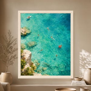 Framed aerial coastal wall art showing swimmers scattered through crystal-clear turquoise water beside a rocky Mediterranean-style cove with a small white boat and bright red swim ring.