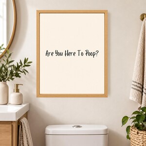 May include: A rectangular wall art piece featuring a light beige background and a thin wooden frame. The text "Are You Here To Poop?" is printed in black, sans-serif lettering in the center of the print, creating a clear and legible display.