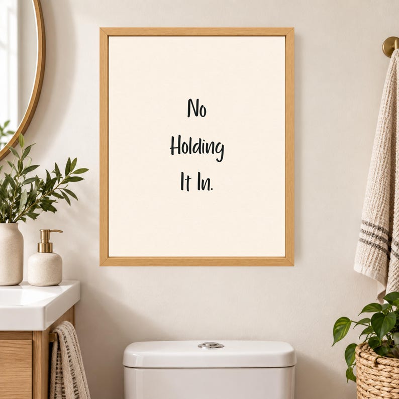 May include: A rectangular wall art print in a light wood frame featuring a cream-colored background with the black text No Holding It In centered in the middle. The print is displayed on a wall above a white toilet tank in a bathroom setting.