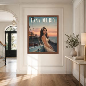 May include: A framed wall art print featuring a portrait of Lana Del Rey wearing a light-colored dress and a headband, leaning against a light blue convertible car by the ocean at sunset, with the text LANA DEL REY printed at the top.