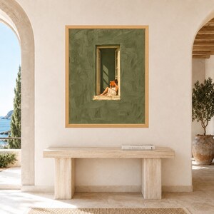 May include: A framed wall art print featuring a textured olive green wall with a central open window. A person sits on the window ledge reading an orange book. The artwork is displayed in a light wood frame, hanging on a plain off-white wall.