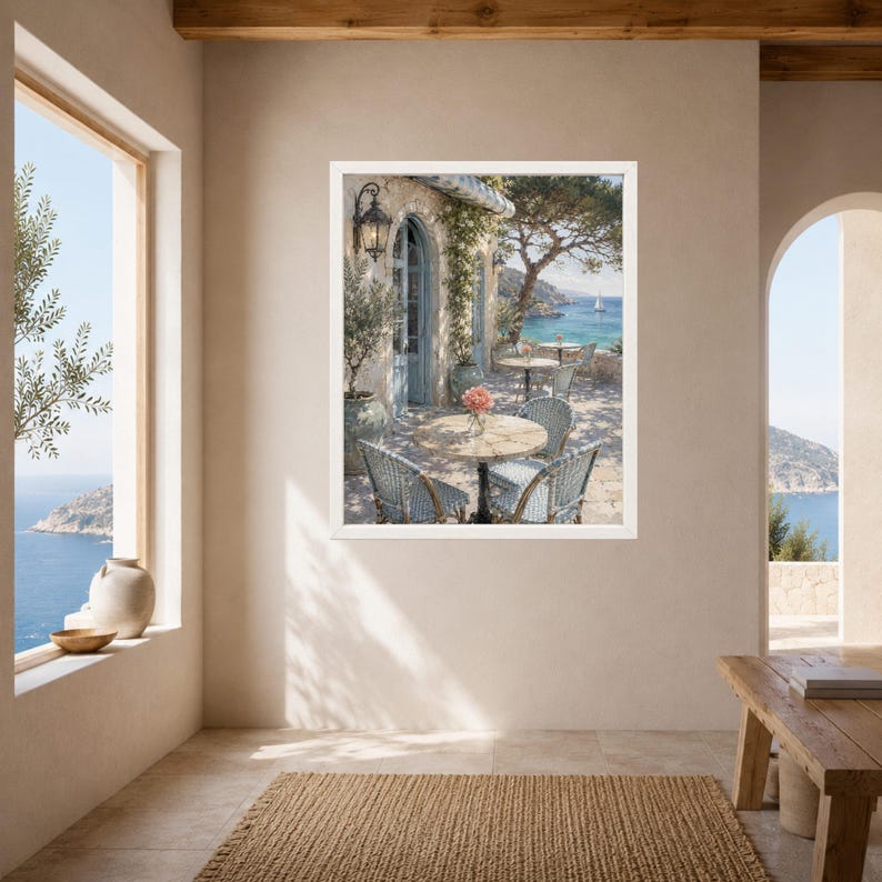 May include: A framed wall art print depicting a Mediterranean coastal terrace with a round stone table, light blue woven chairs, and a vase of pink flowers. The scene features a stone building, a wall lantern, and a view of the blue sea with a sailboat.