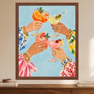 May include: A framed art print depicting four hands raised in a toast, each holding a different cocktail. The drinks include an orange spritz, a martini with olives, a grapefruit drink, and a pink cocktail with cherries, all against a light blue background.