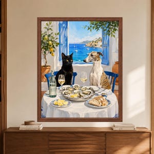 Framed coastal animal wall art showing a black cat and brindle greyhound seated at a Mediterranean dining table with oysters, white wine, lemons and bread overlooking a bright blue sea through open shutters.