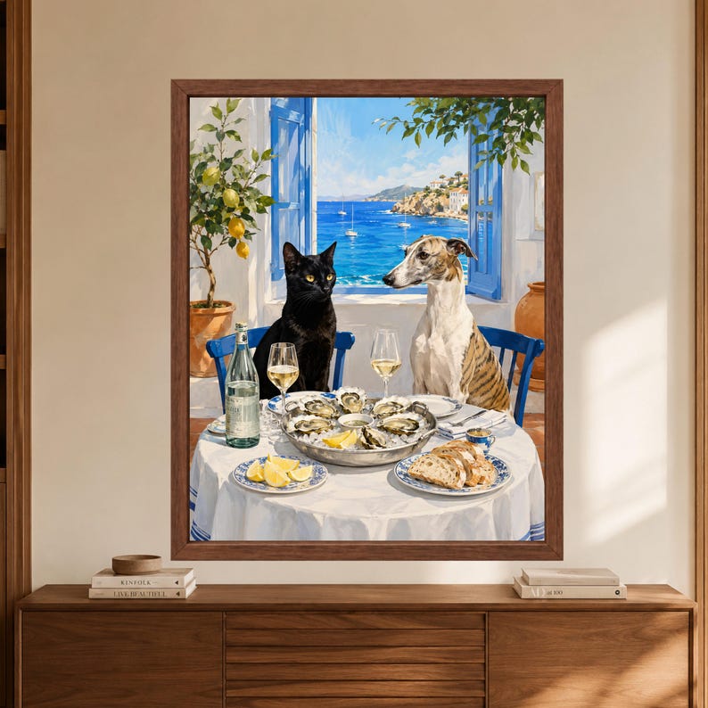 Framed coastal animal wall art showing a black cat and brindle greyhound seated at a Mediterranean dining table with oysters, white wine, lemons and bread overlooking a bright blue sea through open shutters.