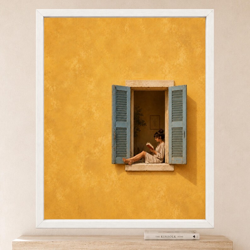 Puede incluir: Una impresi&oacute;n de arte mural enmarcada que muestra una pared de color amarillo mostaza con textura y una ventana abierta. La ventana tiene contraventanas azules de lamas y un alf&eacute;izar de piedra. Una persona sentada lee un libro.