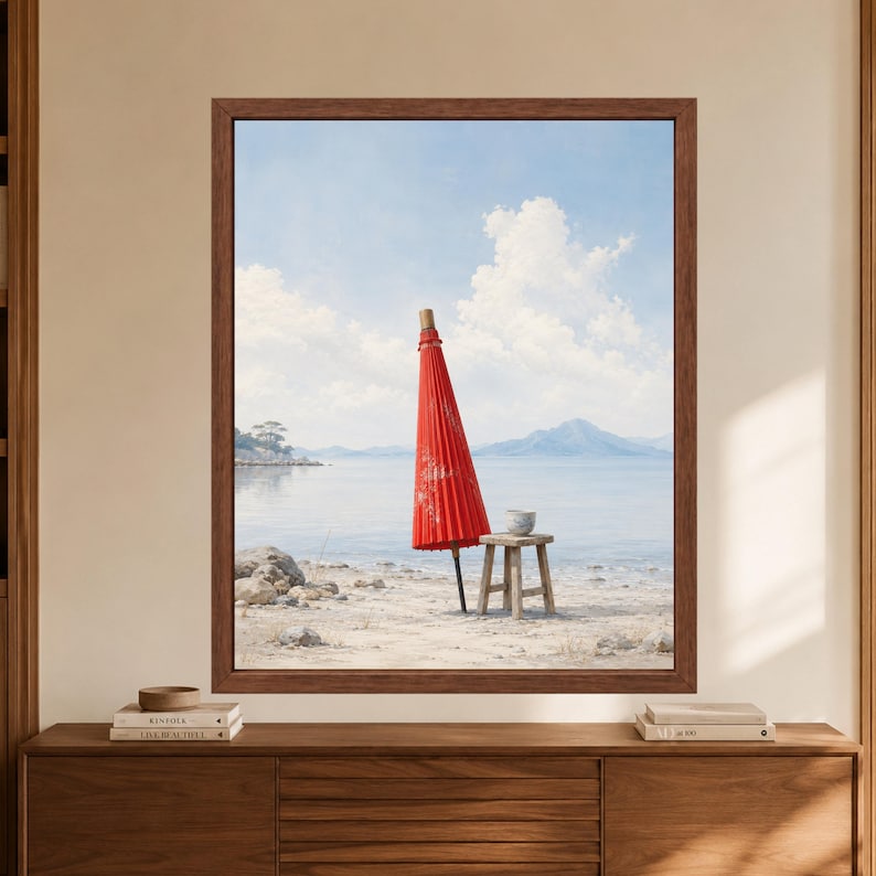 Framed Japanese red umbrella wall art showing a closed red wagasa beside a small wooden stool with a tea cup on a pale beach with calm blue water, distant mountains and soft clouds.
