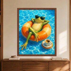 Framed funny frog wall art showing a green frog wearing dark sunglasses and relaxing cross-legged on an orange pool float beside a small espresso cup in bright turquoise swimming pool water.