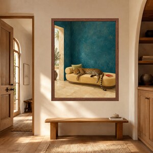 May include: A framed wall art print featuring a leopard resting on a long, curved, beige upholstered bench. The scene is set against a textured teal wall with a view of a bright outdoor space. A small red book lies on the bench near the leopard.