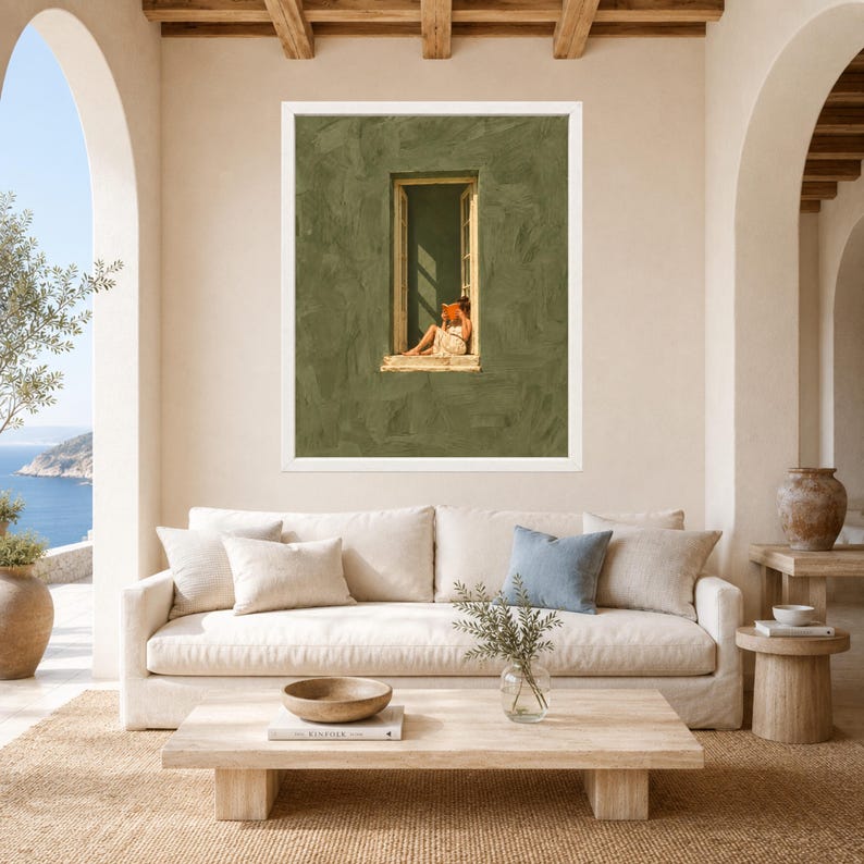May include: A framed wall art print featuring a textured olive green wall with a central open window. A person sits in the window frame reading an orange book while wearing a light-colored dress. The artwork is displayed in a white frame.