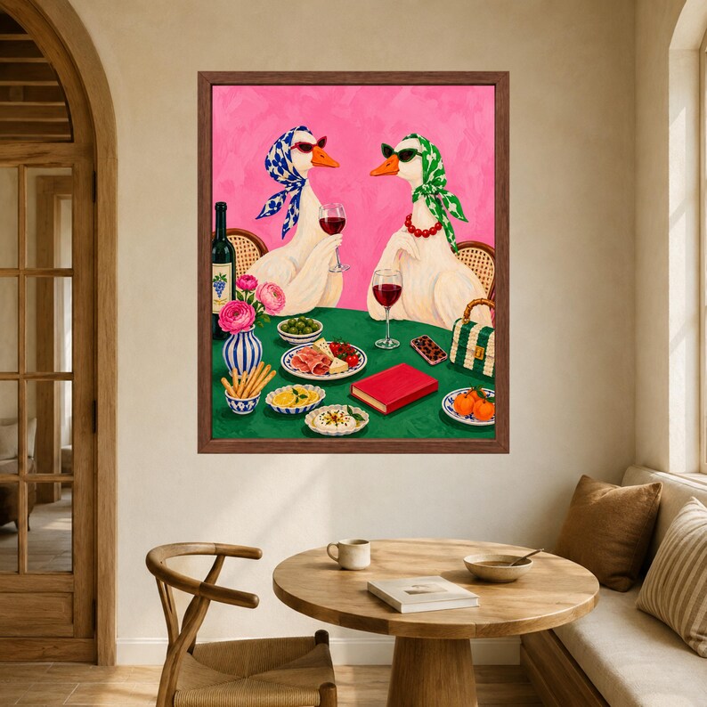 May include: A framed wall art print featuring two white geese wearing headscarves and sunglasses, sitting at a green table with wine, food, and a red book. The background is a vibrant pink, and the geese are positioned as if dining together.