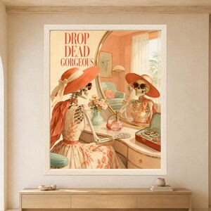 Framed Drop Dead Gorgeous skeleton wall art featuring a stylish skeleton wearing a coral sun hat and sunglasses while sitting at a retro vanity with perfume, jewelry and mirror reflection.