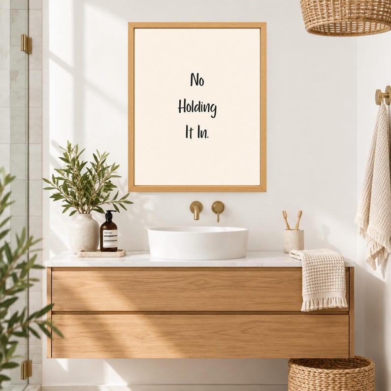 May include: A rectangular wooden frame containing a cream-colored print with the black text "No Holding It In." The frame is mounted on a white wall above a white vessel sink, a wooden vanity, a potted plant, and a hanging towel in a bathroom.