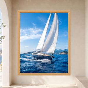 Op de afbeelding: Een ingelijste wandkunstprint met een donkerblauw zeiljacht met witte zeilen dat door diepblauw oceaanwater vaart onder een heldere lucht met lichte wolken, met in de verte rotsachtige eilanden zichtbaar op de horizonlijn.